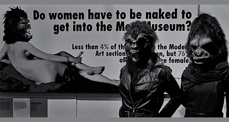 Due componenti delle Guerrilla Girls, con le caratteristiche maschere da gorilla, davanti al manifesto Do Women Have to Be Naked to Get into the Met. Museum? (1989), esposto al Victoria and Albert Museum di Londra. Fotografia del 2014. Foto: Eric Huybrechts / Wikimedia Commons — CC BY-SA 2.0